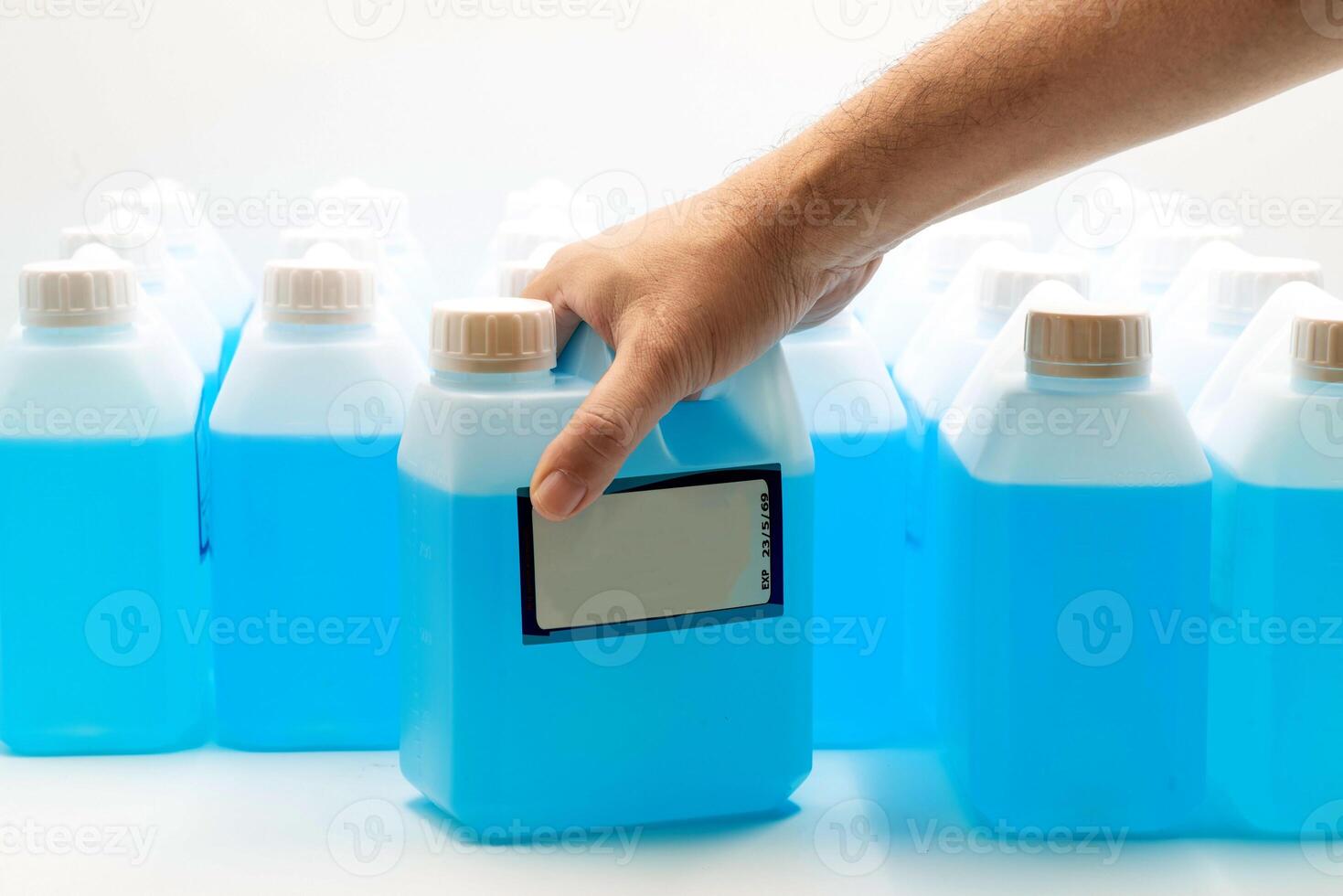 Hand holding a container of ethyl alcohol among multiple blue liquid bottles in a storage area photo