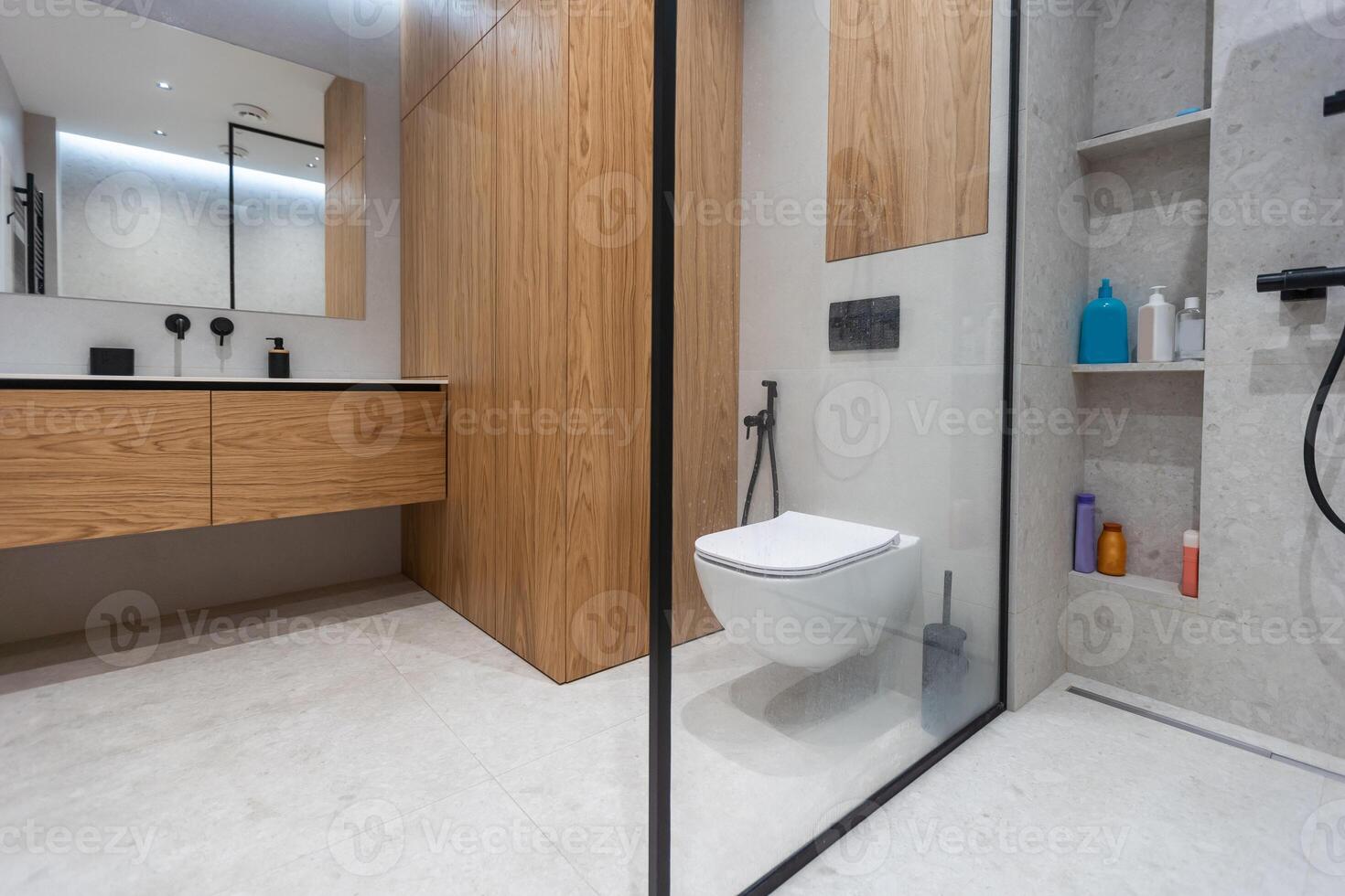 Bathroom in a modern style with gray and white tiles. There is a large mirror with luminous lamps, tabletop with wooden drawers and sink, bath with shower and glass partition, towel rack and a hanger. photo