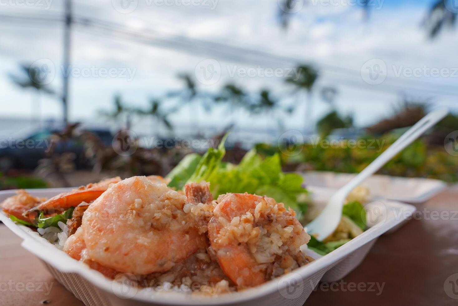 A plate of garlic shrimp over rice with greens on a wooden table, set against palm trees, ocean views, parked cars, and a blue sky in Hawaii. photo