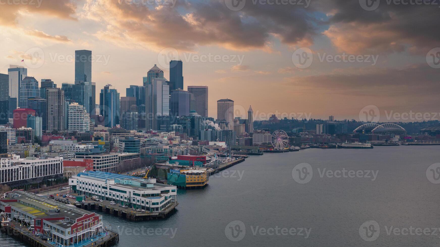 Downtown Seattle's skyline features the Space Needle, skyscrapers, a cargo ship in the bay, the Seattle Great Wheel, and Lumen Field at sunset. photo