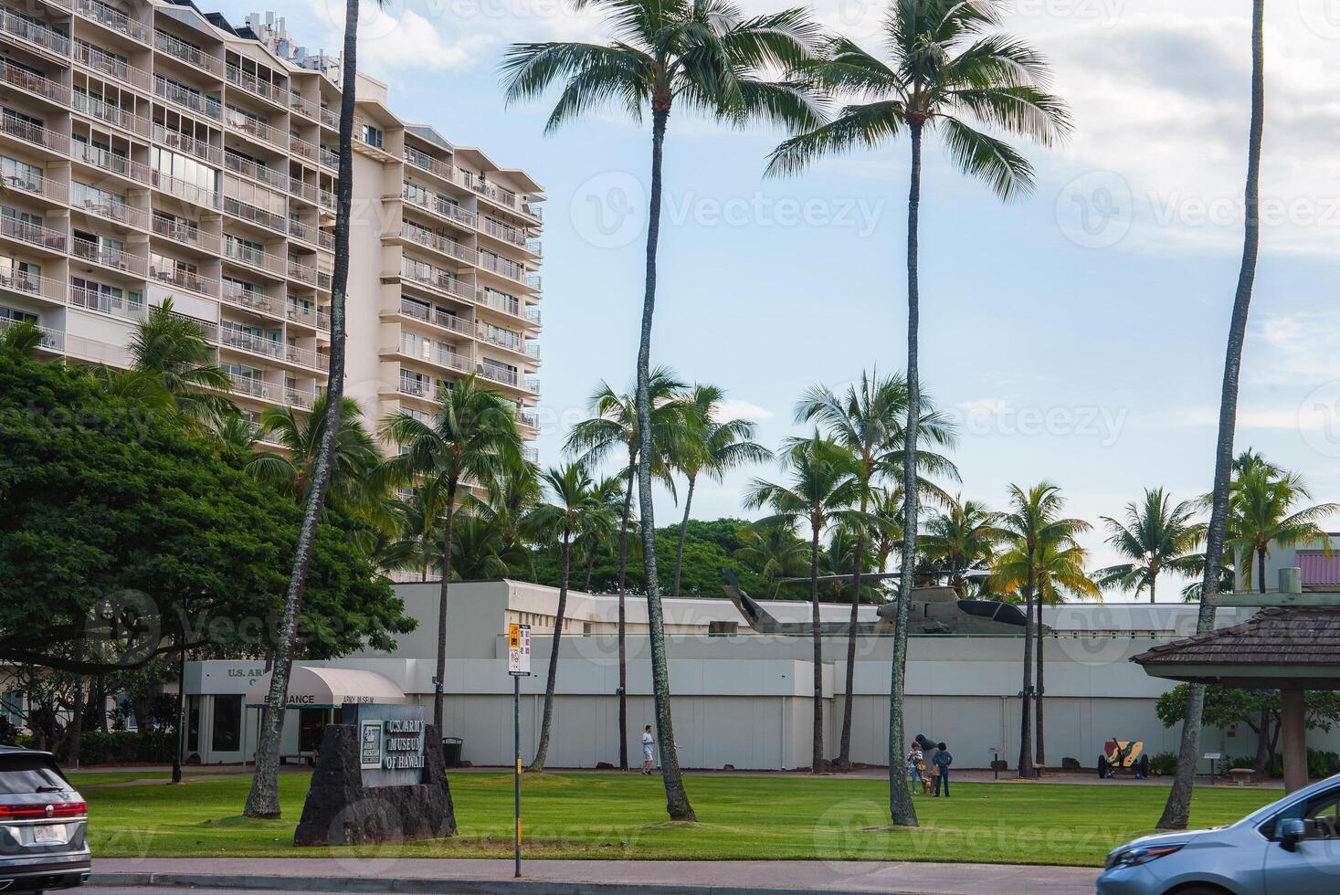 A grassy area with tall palm trees, a modern building, and the U.S. Army Museum of Hawaii sign. People walk and relax under sunny, clear skies. photo