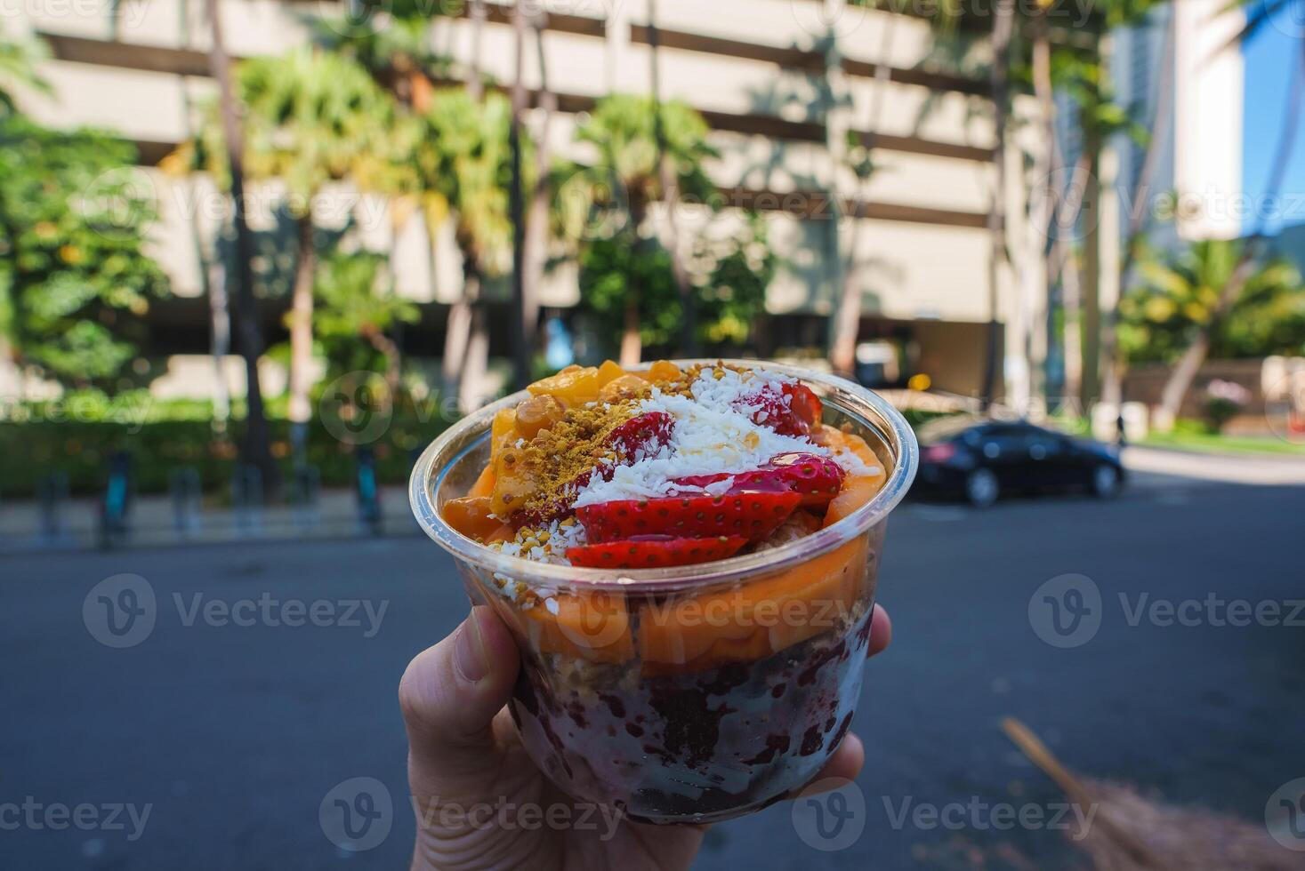 A colorful acai bowl with strawberries, mango, coconut, and granola is held against a modern building, palm trees, and a street in Honolulu. photo