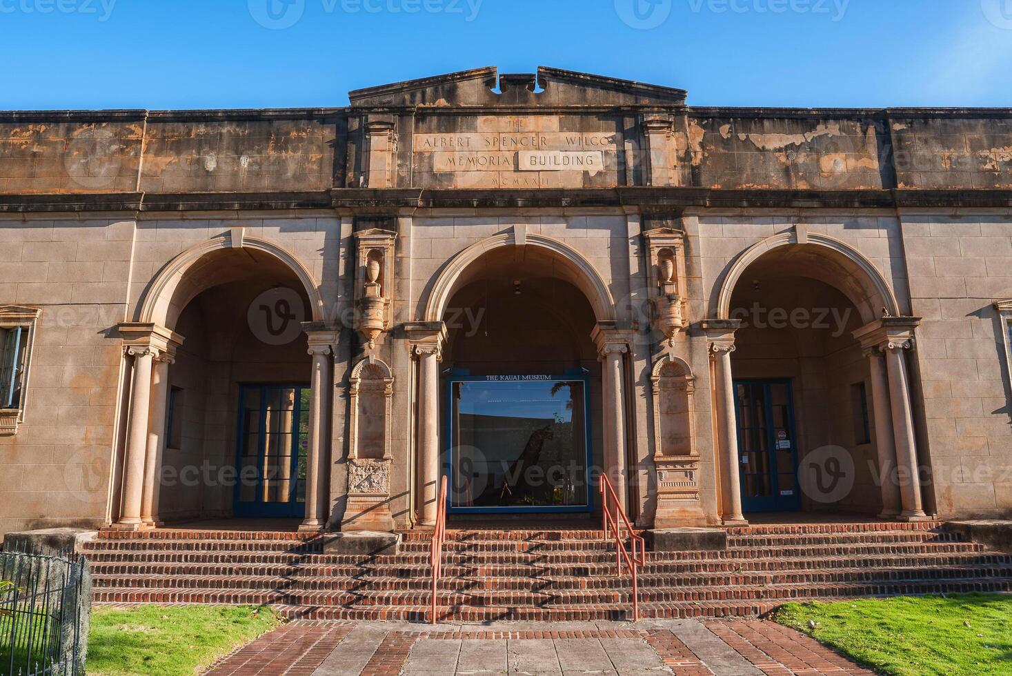 Historic Albert Spencer Wilcox Memorial Building in Hawaii, featuring arched entrances, decorative columns, brick steps, and a small grassy area. photo