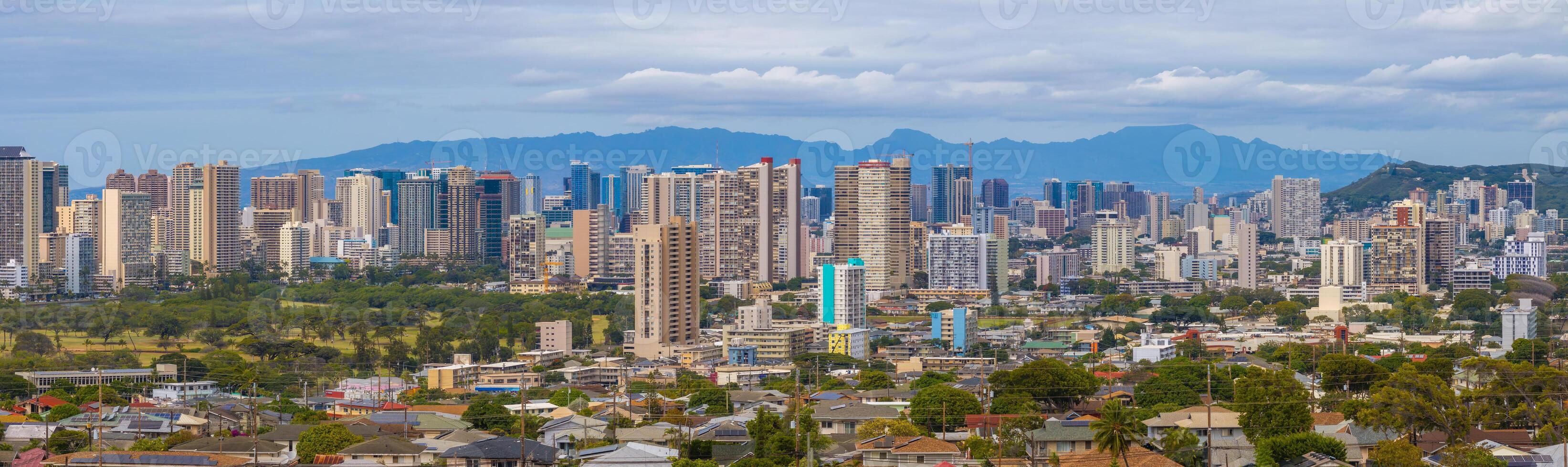 View of Honolulu, Hawaii, featuring high rise buildings, residential areas, green spaces, and the Ko olau Mountain Range under an overcast sky. photo