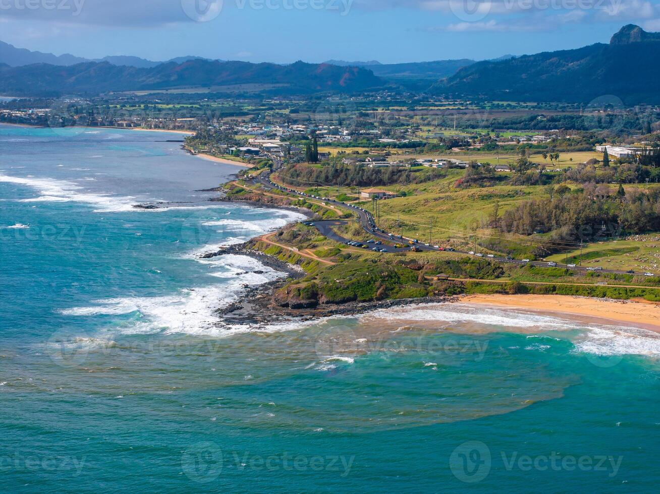Aerial image of Kauai, Hawaii, showing turquoise waters, sandy beaches, a winding coastal road, green fields, and distant mountains under a cloudy sky. photo