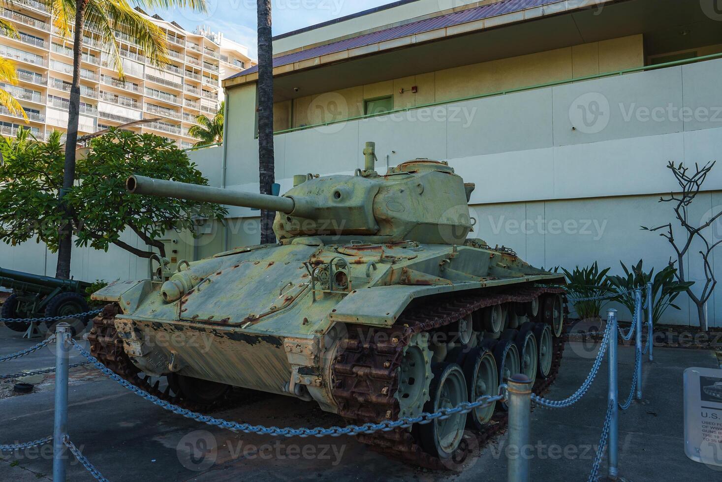 A weathered green military tank with rusted tracks is displayed outdoors near a light colored building, palm trees, and a multi story structure in Hawaii. photo