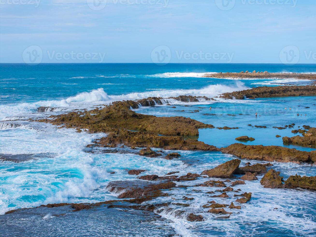 Rugged rocky formations surrounded by vibrant blue ocean waters on Oahu island. Waves crash, creating foam, with people visible near calmer pools. photo
