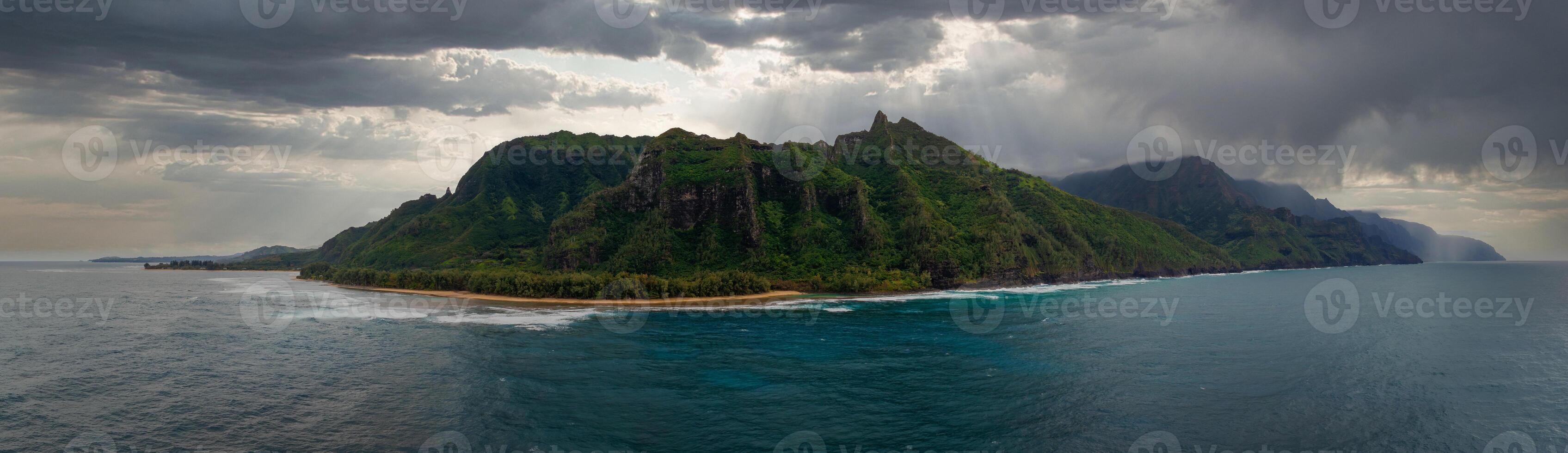 Lush green cliffs of the Napali Coast rise from the ocean, with a sandy beach and palm trees along the shoreline under a cloudy, sunlit sky. photo