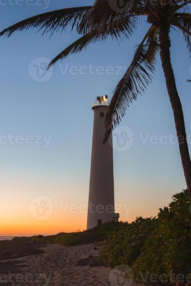 A tall white lighthouse stands against a sunset sky, framed by a palm tree and lush greenery, with a sandy path leading to its base in Hawaii. photo