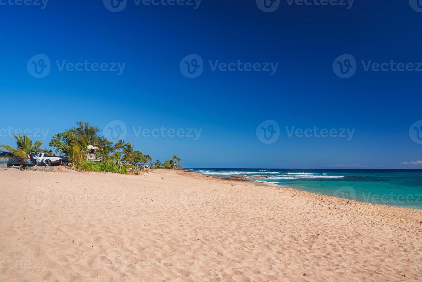 A tranquil beach scene with golden sand, turquoise waters, palm trees, and small structures, including a parked vehicle, under a vibrant blue sky. photo