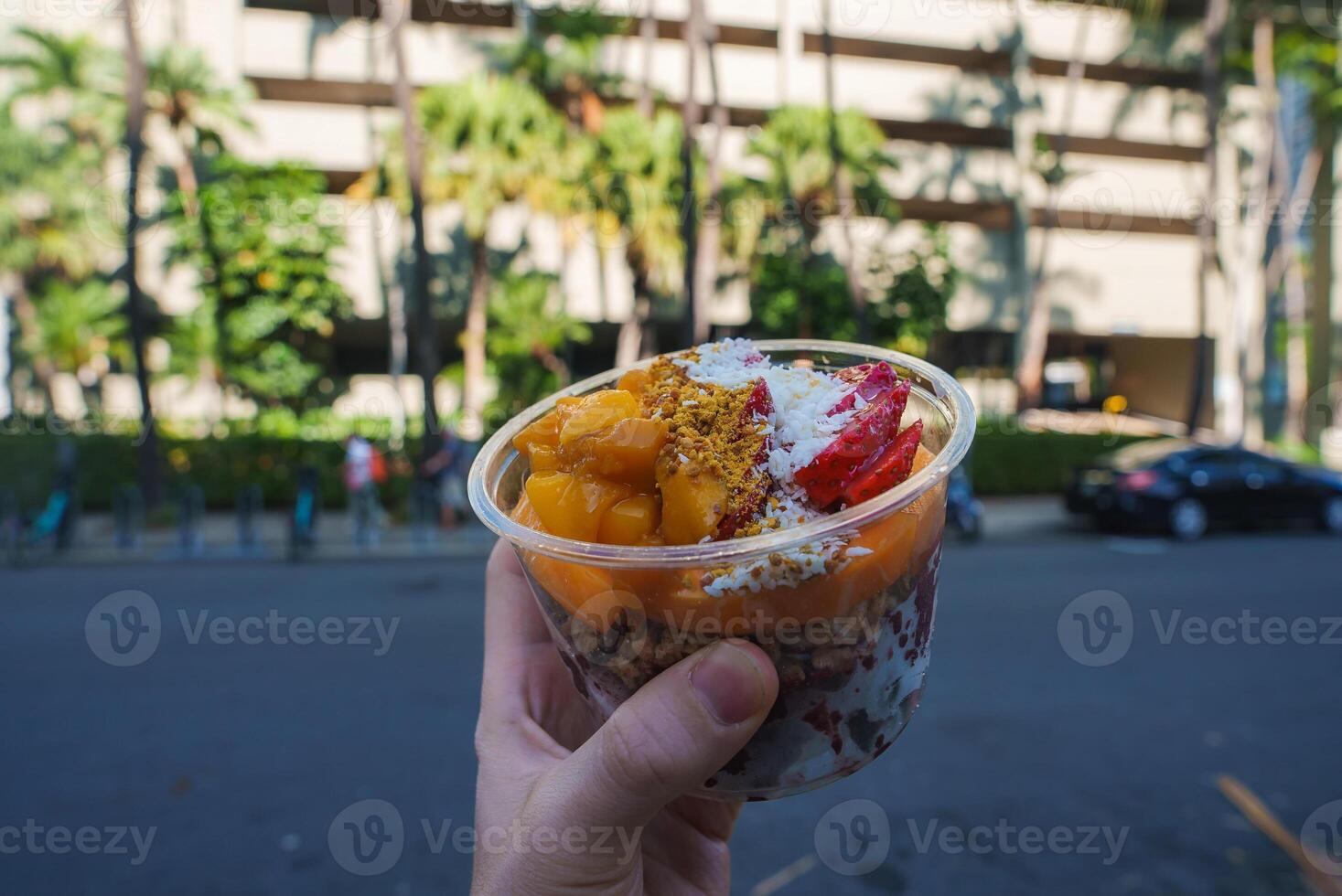 A clear plastic bowl with fresh fruits, granola, and coconut shavings held against a street with palm trees and a multi story building in Honolulu. photo