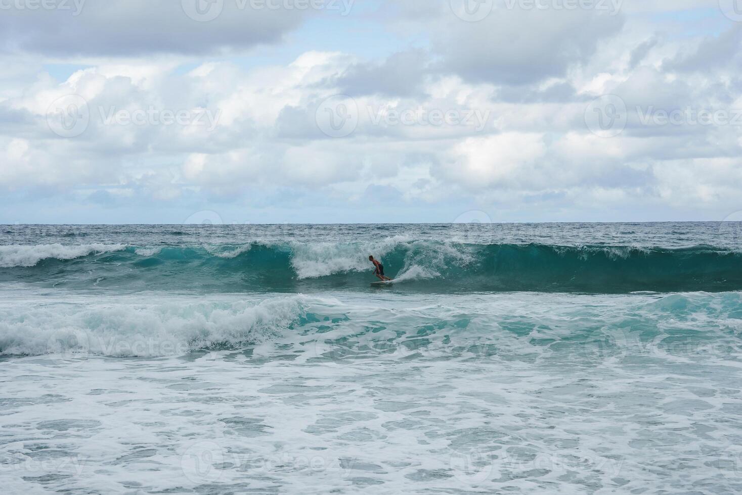 A lone surfer navigates a curling wave in turquoise waters under a sky with scattered clouds, showcasing the energy of the ocean in Hawaii. photo