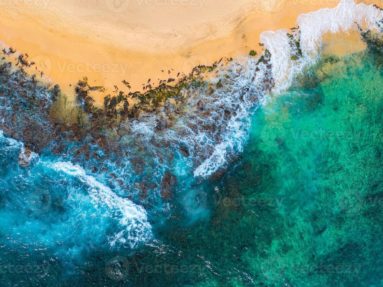 Aerial perspective of Oahu island's coastline in Hawaii, showing a sandy beach, turquoise waters, rocky formations, and green vegetation along the shore. photo