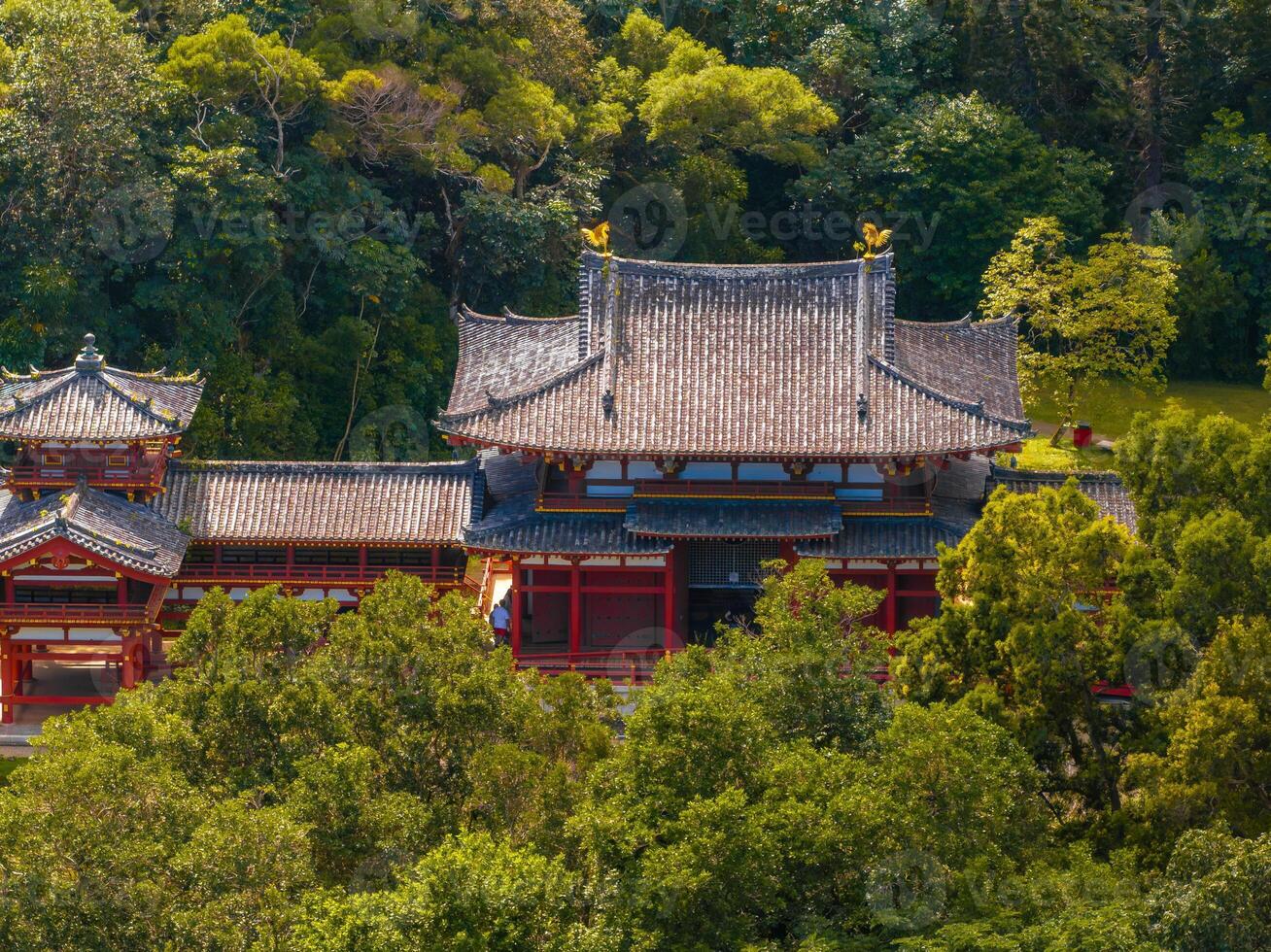 The Byodo In Temple in Oahu, Hawaii, features red wooden structures, a curved tiled roof, and golden accents, set amidst dense trees and greenery. photo