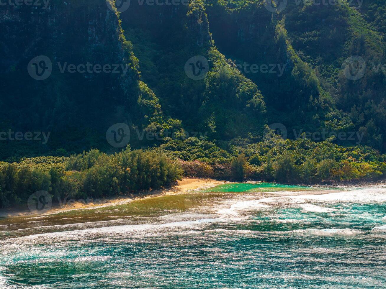 A coastal scene on Kauai island with a sandy beach, turquoise ocean waves, lush greenery, and dramatic Napali cliffs covered in dense vegetation. photo