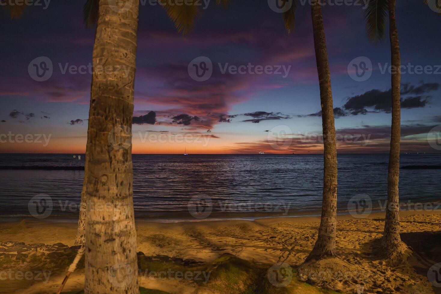 A tranquil beach scene at sunset with vibrant orange, pink, and purple skies. Palm trees frame the view, and small boats float on the calm ocean. photo