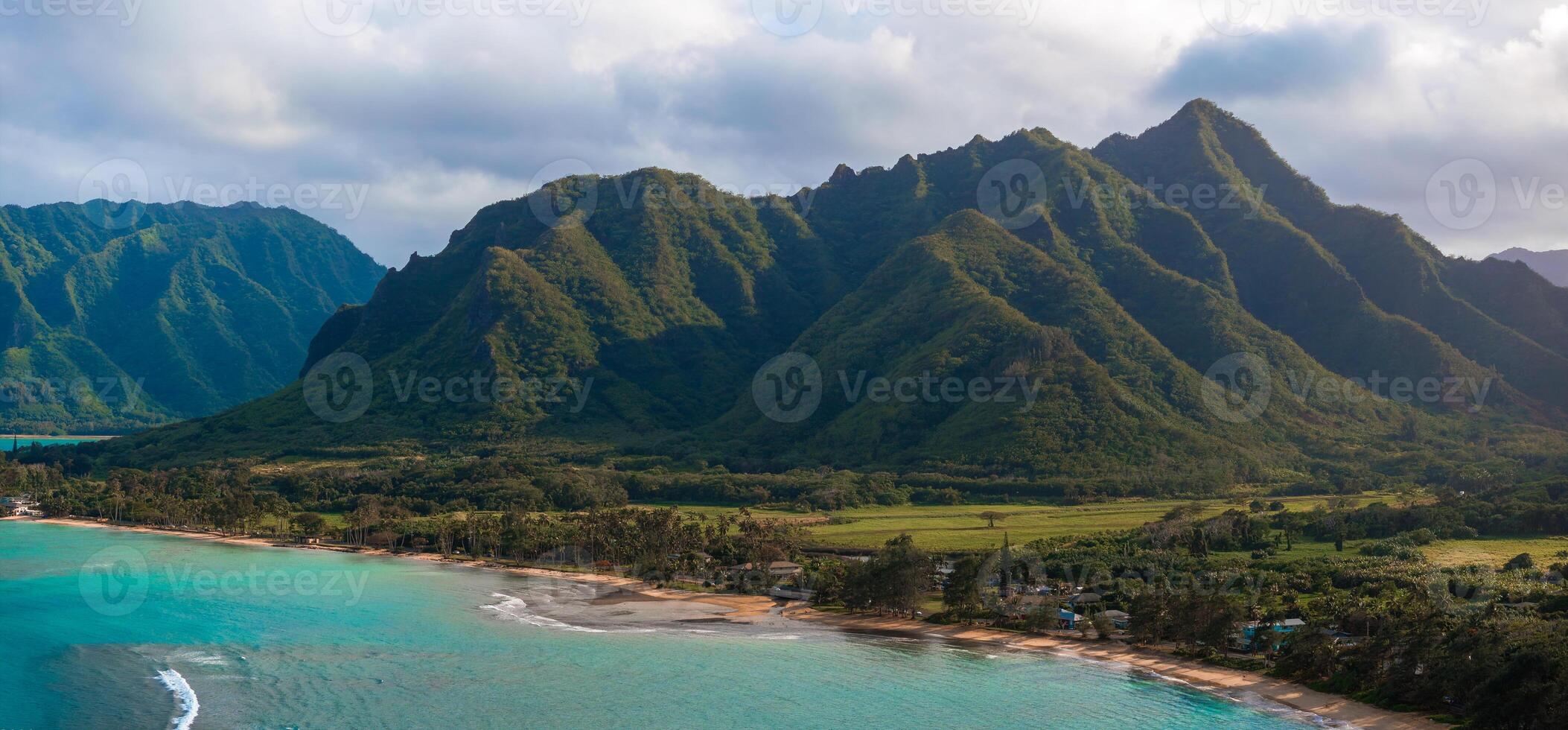 Aerial view of Oahu, Hawaii, showing green mountains with sharp ridges, a turquoise shoreline, sandy beach, small settlement, and dense vegetation. photo