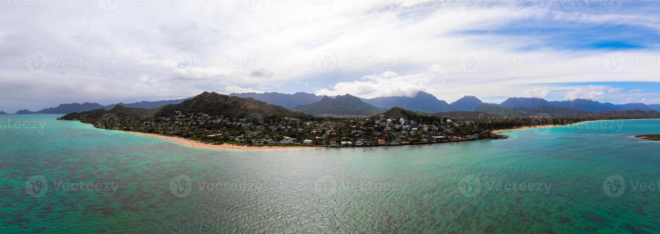 Aerial view of Oahu, Hawaii, showing a sandy beach, turquoise waters, coral reefs, residential homes, and lush green mountains under a cloudy sky. photo