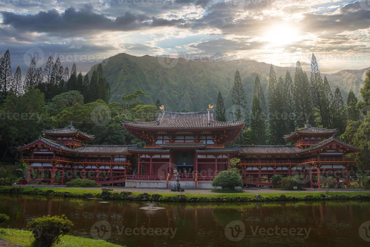 The Byodo In Temple stands amidst lush greenery, a reflective pond, and the Ko olau mountain range under a partly cloudy sky in Oahu, Hawaii. photo