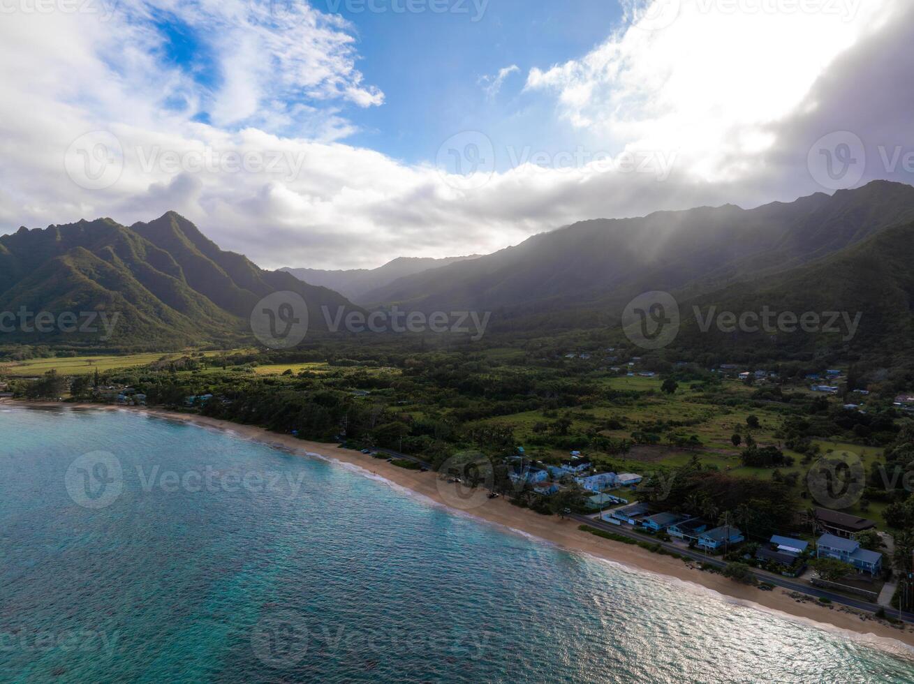 Aerial view of Oahu, Hawaii, showing turquoise waters, a sandy beach, a small community, and lush green mountains under a partly cloudy sky. photo