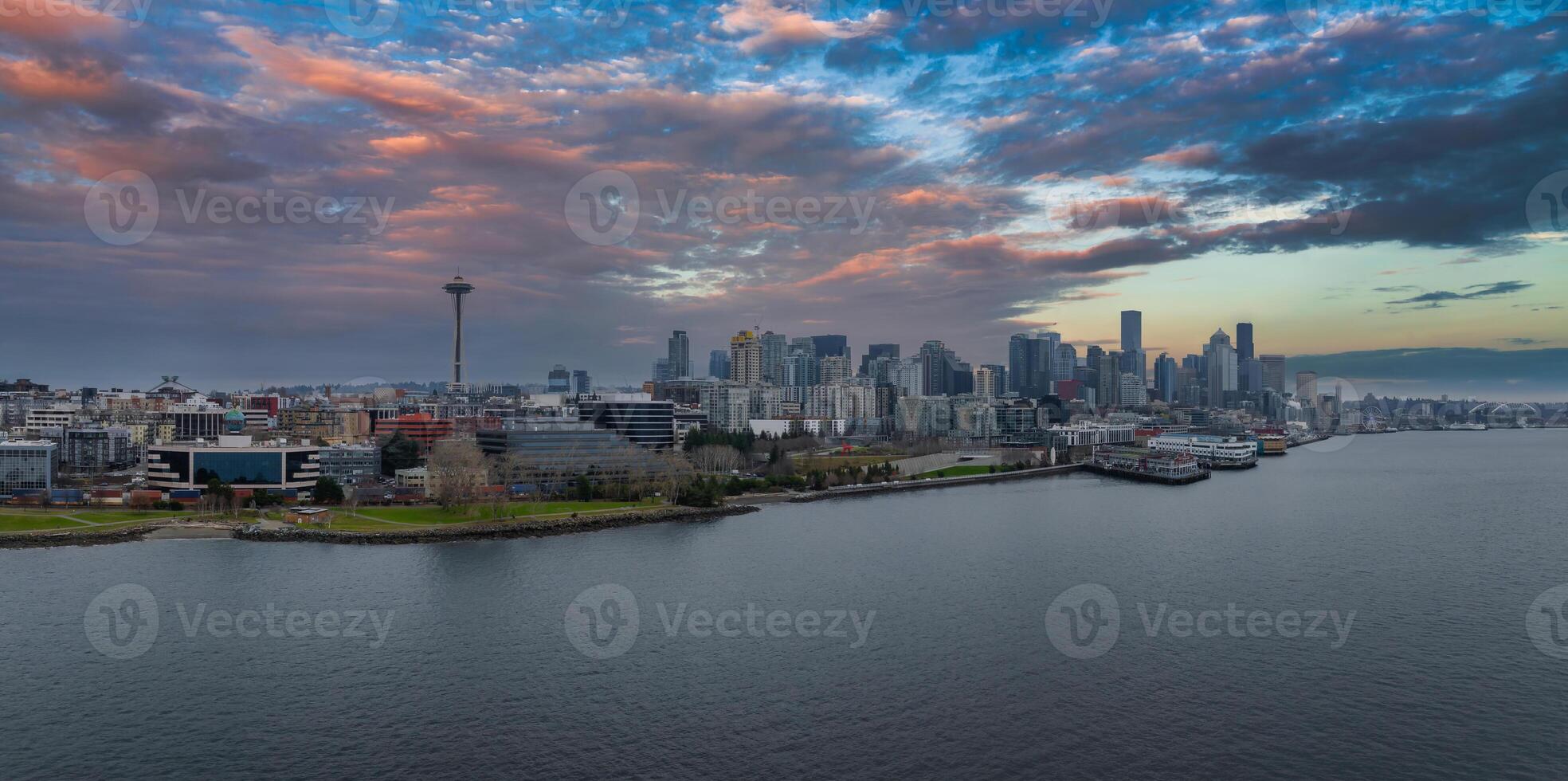 Downtown Seattle skyline with the Space Needle, modern skyscrapers, calm waterfront, and a cargo ship in the bay under a colorful sunset sky. photo