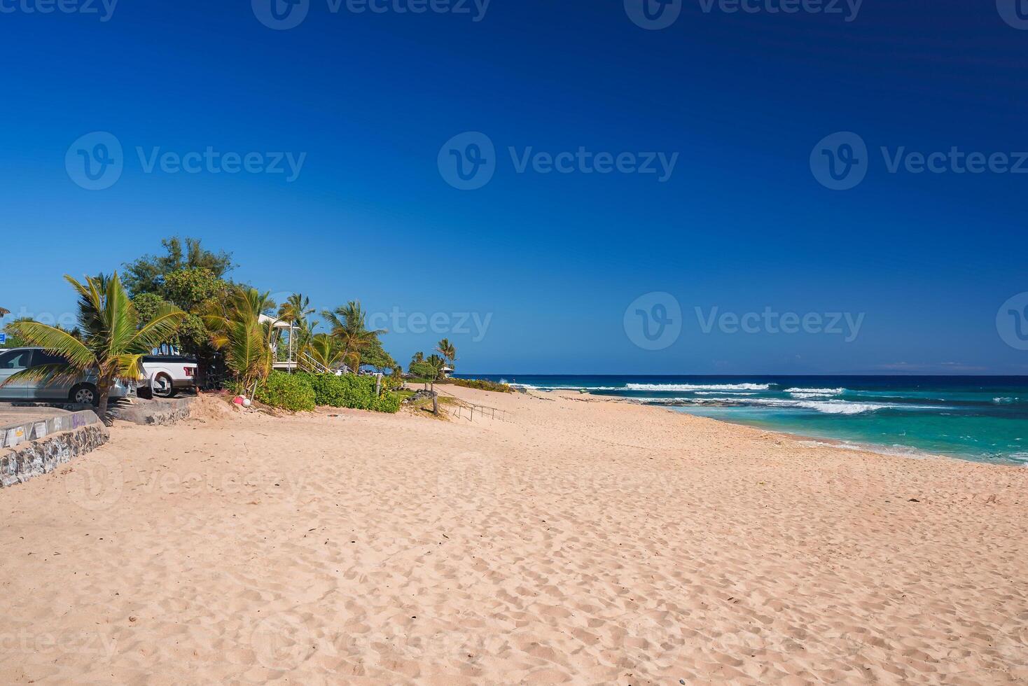 A tranquil sandy beach with turquoise waves, palm trees, and lush greenery under a clear blue sky. Parked vehicles near a stone wall suggest access nearby. photo