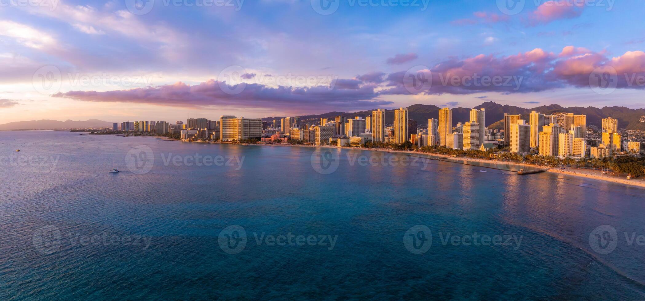 Aerial view of Waikiki Beach in Honolulu, Oahu, Hawaii, at sunset. High rise buildings line the shore, with mountains and colorful skies in the background. photo