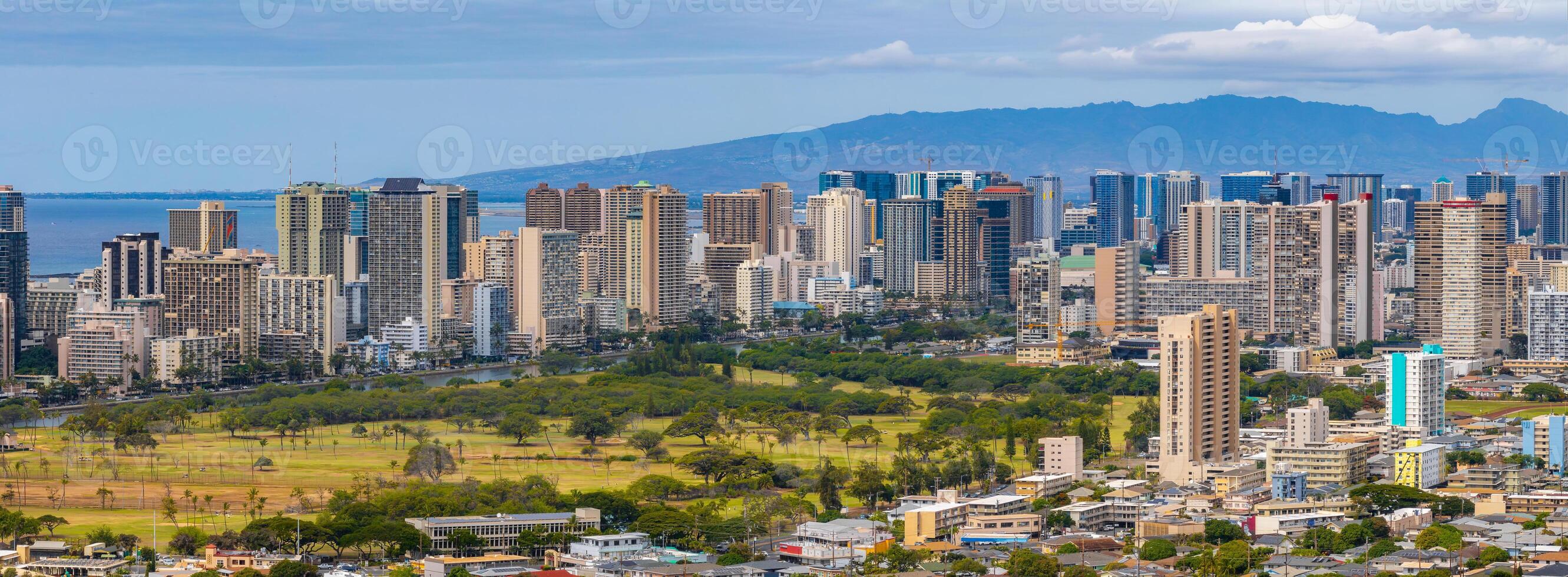 Aerial view of Honolulu, Hawaii, featuring modern high rise buildings, a green park, the Pacific Ocean, and a distant mountain range under clear skies. photo