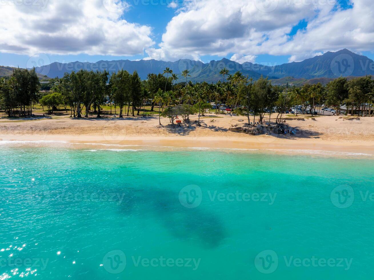 Golden sandy beach on Oahu island, Hawaii, with turquoise waters, palm trees, a small park, and the Ko olau mountain range under a cloudy sky. photo