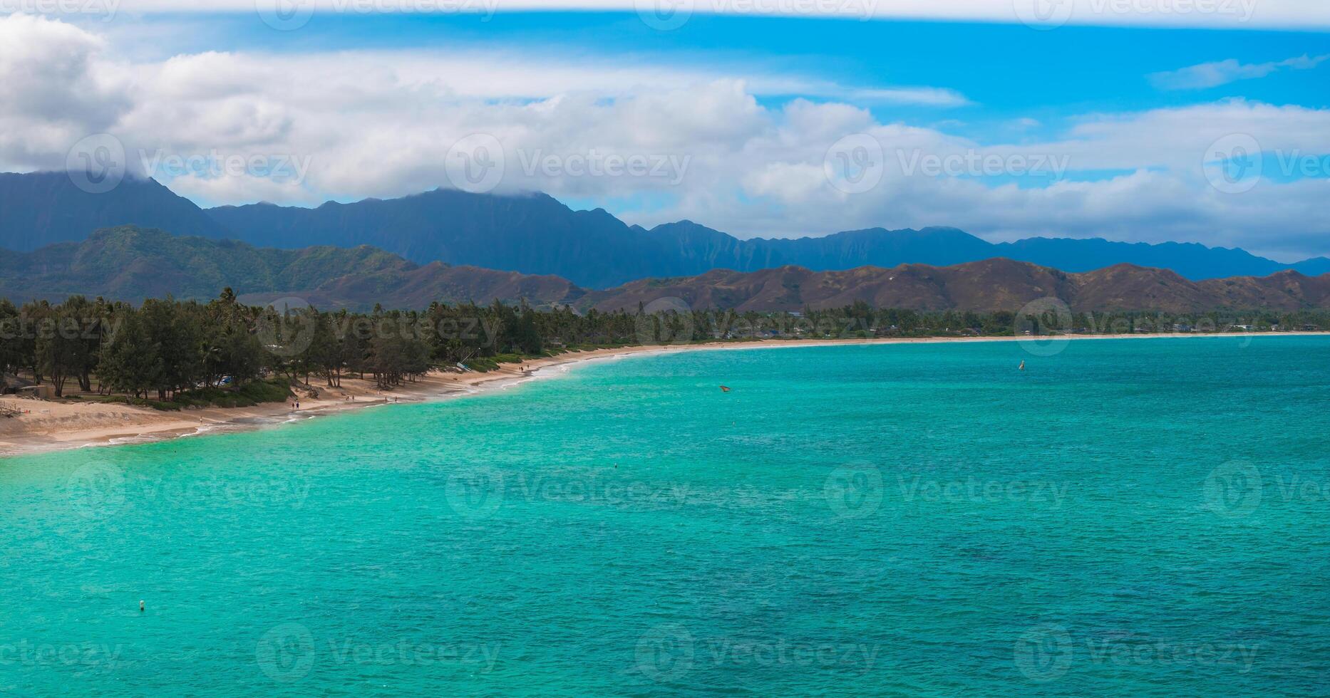 Aerial view of a sandy beach with turquoise waters on Oahu island, Hawaii, bordered by trees and backed by lush green and brown mountains under a cloudy sky. photo