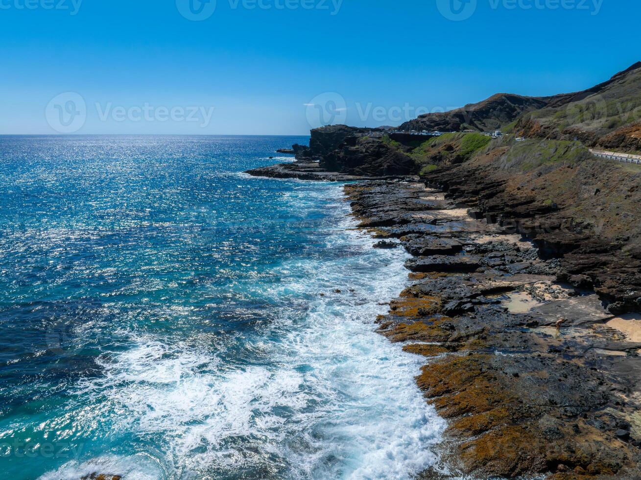 Rugged cliffs and rocky shores meet vibrant blue ocean waves in Oahu, Hawaii. A winding road runs along the hillside under a clear sky. photo