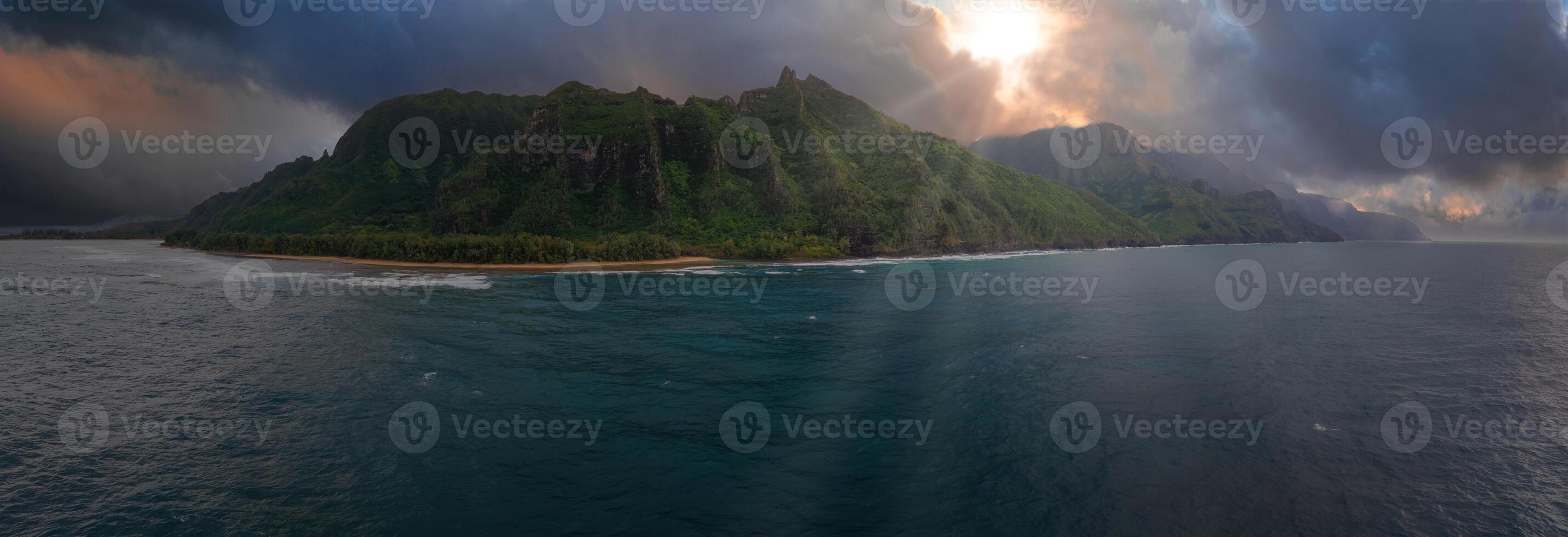 Aerial view of lush green cliffs, sandy beaches, and dramatic clouds along the Napali Coast on Kauai Island, Hawaii, with sunlight breaking through. photo