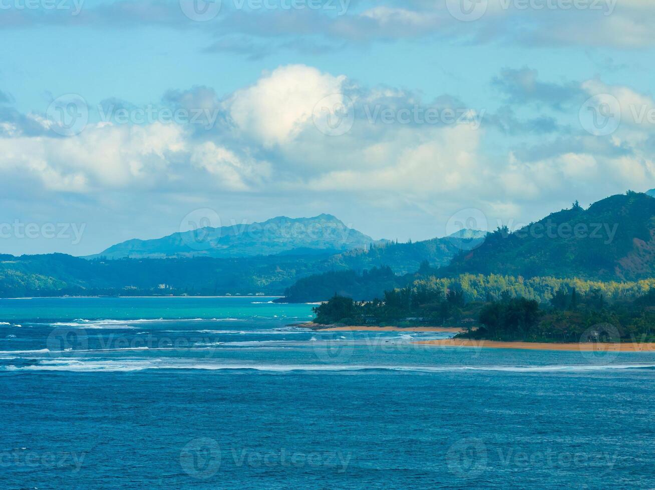 Aerial view of Kauai island's coastline showing turquoise ocean, sandy beach, lush green vegetation, and rolling hills under a partly cloudy sky. photo