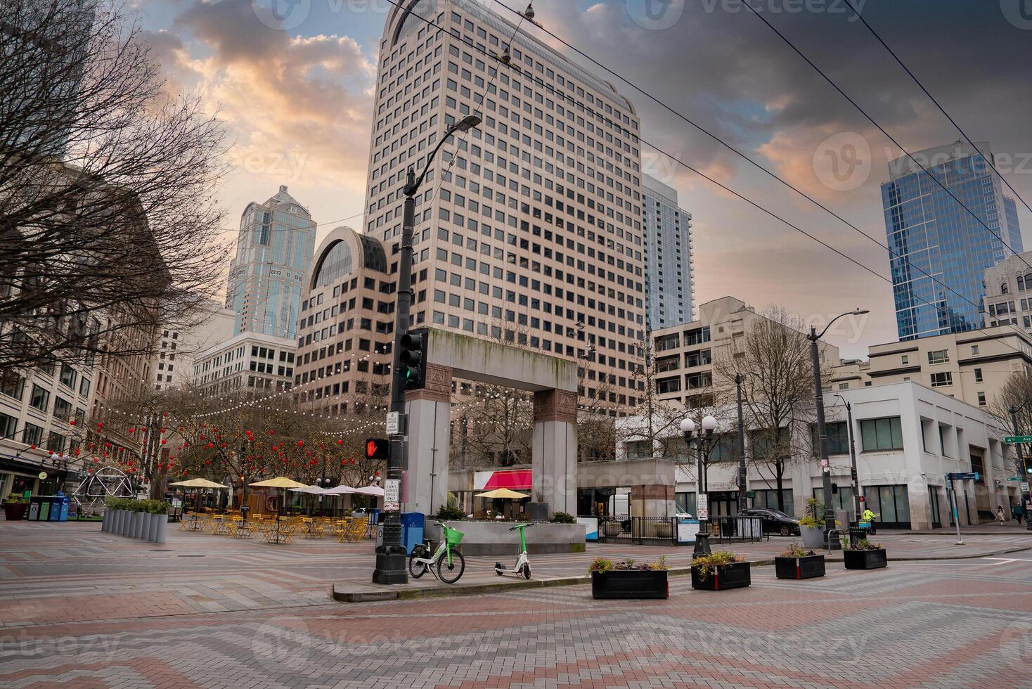 A city plaza in downtown Seattle features a modern archway, tall office buildings, outdoor seating with umbrellas, and a dramatic cloudy sky. photo