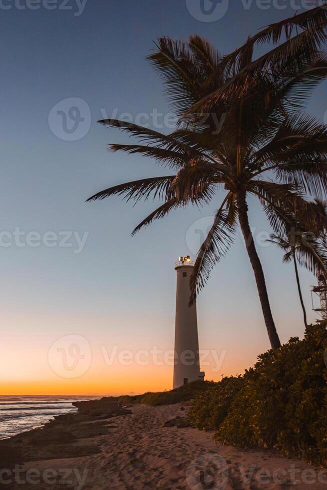 A white lighthouse near the shoreline stands tall at sunset, with a palm tree silhouetted against an orange and blue sky. Sandy beach and greenery surround the scene. photo