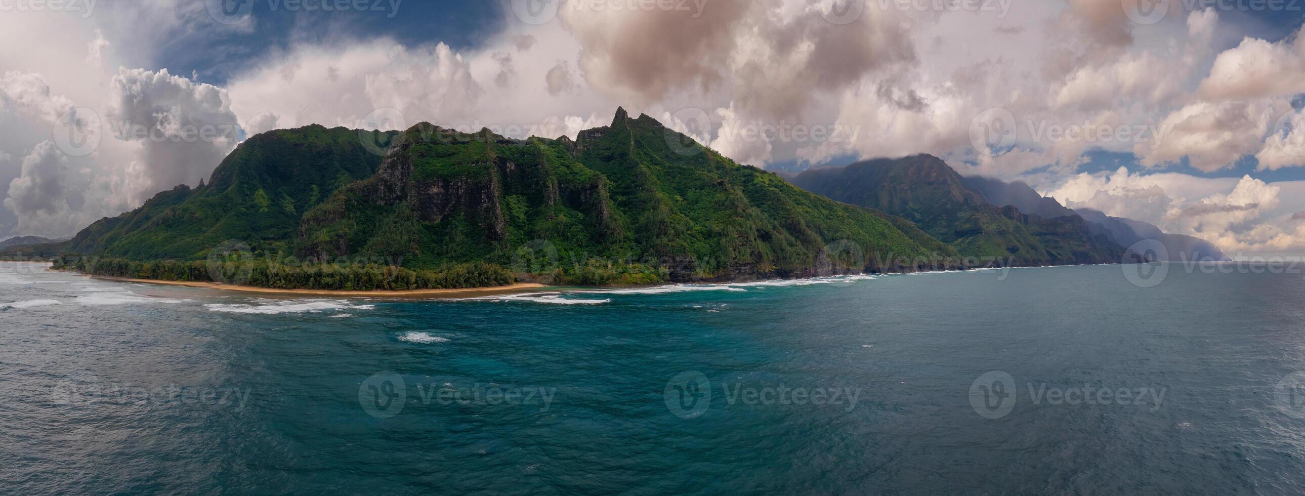 Aerial view of the Napali Coast on Kauai Island, Hawaii, showing rugged green cliffs, a sandy beach, and vibrant blue Pacific Ocean waters under a cloudy sky. photo