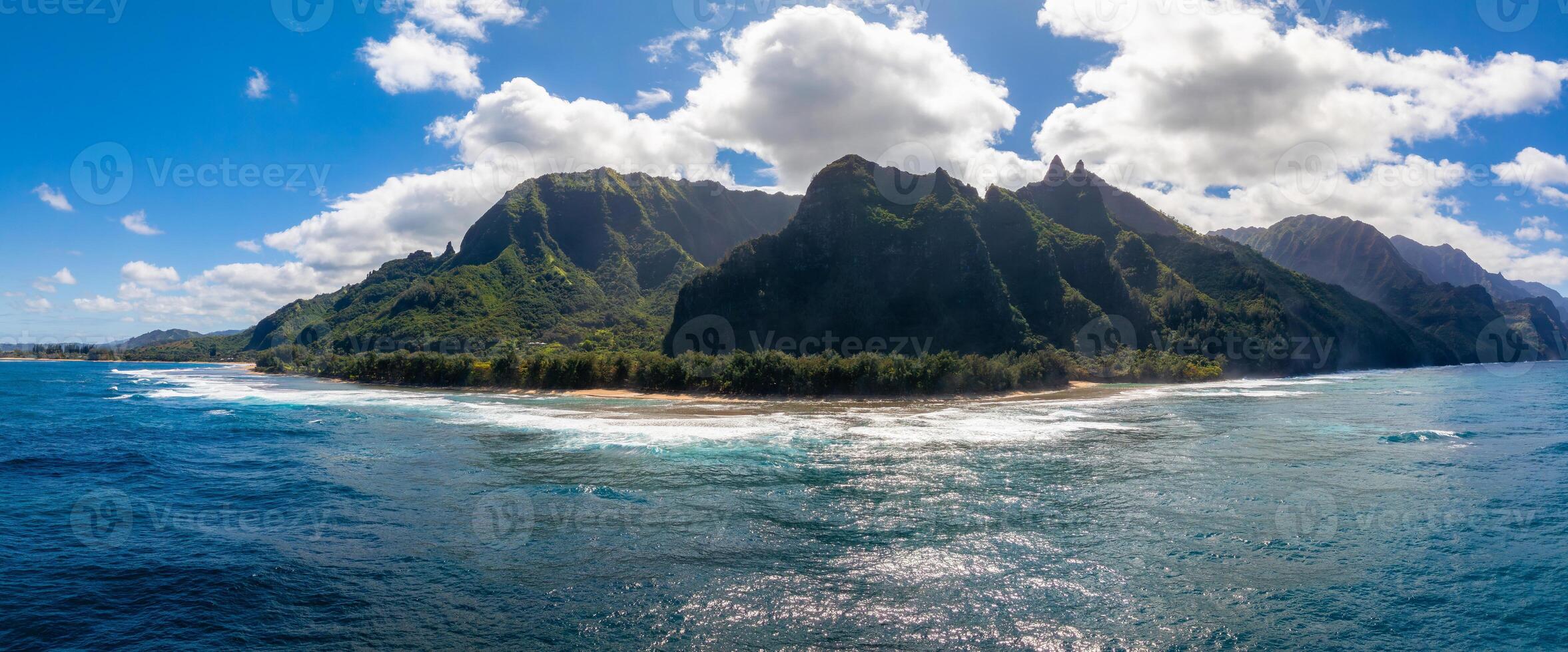 Jagged green cliffs of the Napali Coast rise above a sandy beach and vibrant blue ocean, with lush vegetation and scattered white clouds overhead. photo