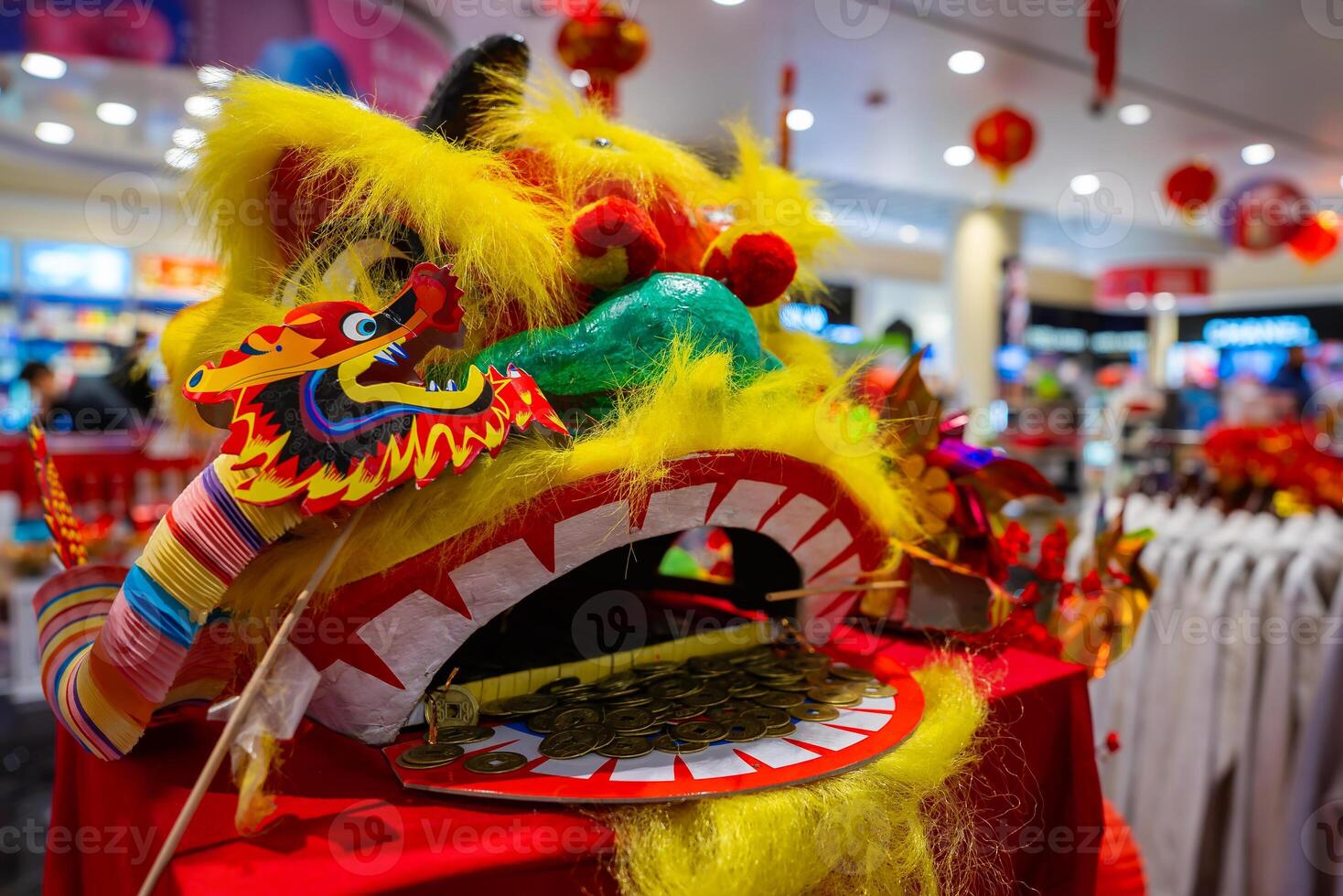 A vibrant Chinese lion dance head with yellow fur and red accents sits on a red table with coins in its mouth. Red lanterns hang in a festive setting. photo