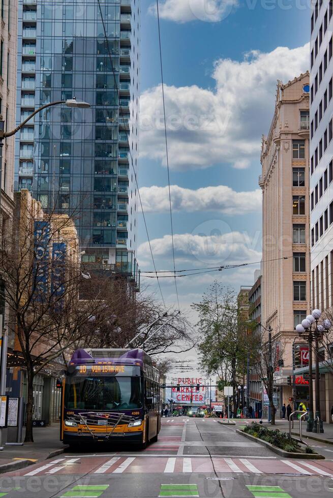 A vibrant street in downtown Seattle, USA, featuring a public transit bus, the iconic Public Market Center sign, modern and historic buildings, and trolley wires. photo