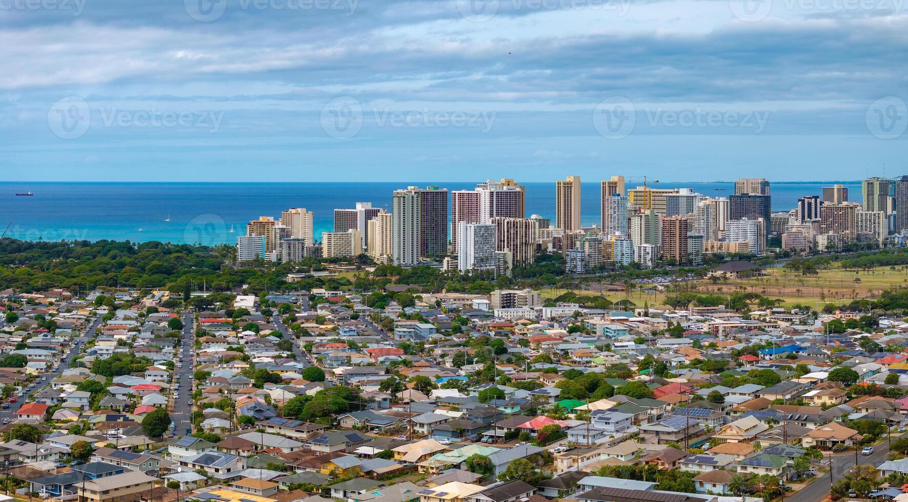 Aerial perspective of Honolulu, Hawaii, featuring high rise buildings, residential areas, calm blue ocean, distant ships, and a partly cloudy sky. photo