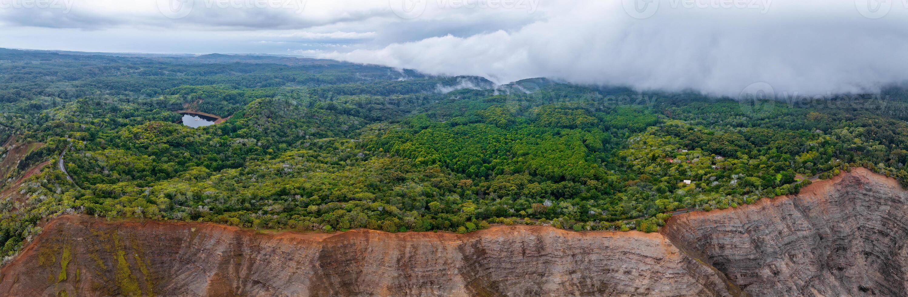 Aerial view of Kauai Island showing rugged reddish cliffs, dense green forest, a small water body, and low lying clouds on the horizon. photo