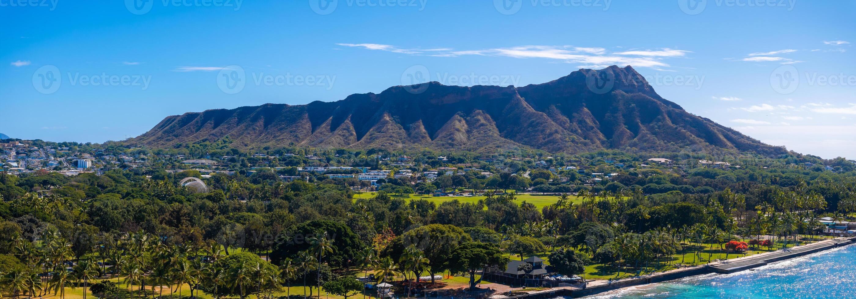 Diamond Head Crater with rugged ridges, lush greenery, palm trees, and a coastal area bordered by a seawall, under a bright blue sky with clouds. photo