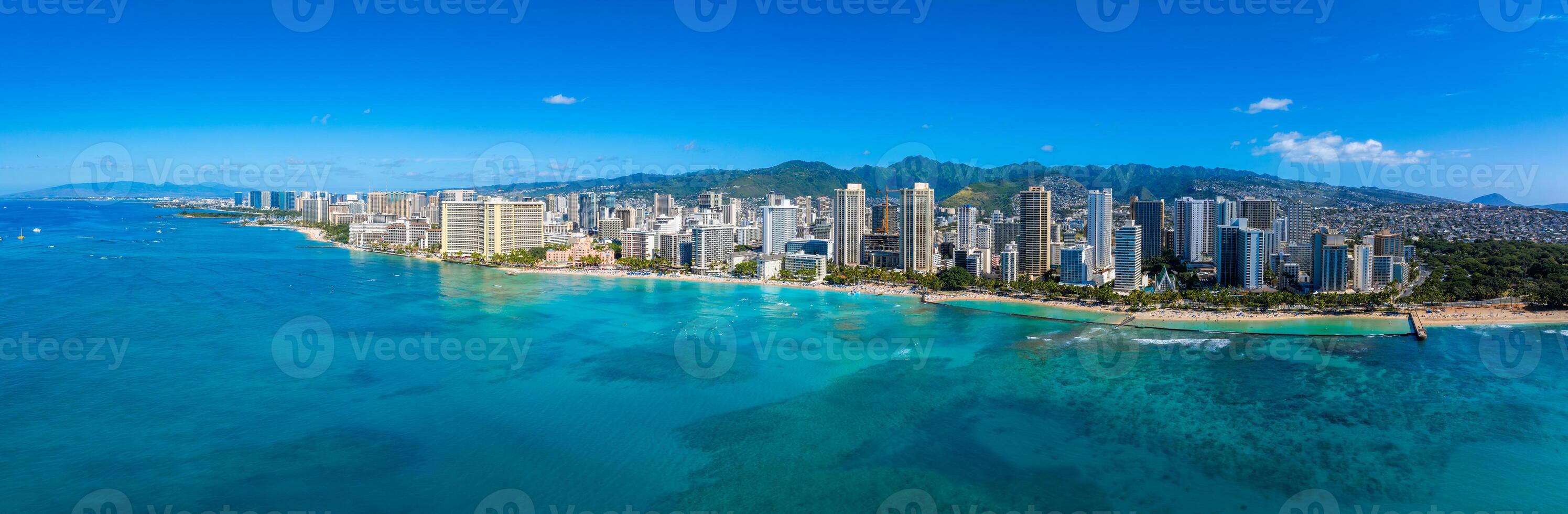 Aerial view of Honolulu, Hawaii, featuring Waikiki Beach, turquoise Pacific waters, high rise buildings, and lush green mountains under a clear sky. photo