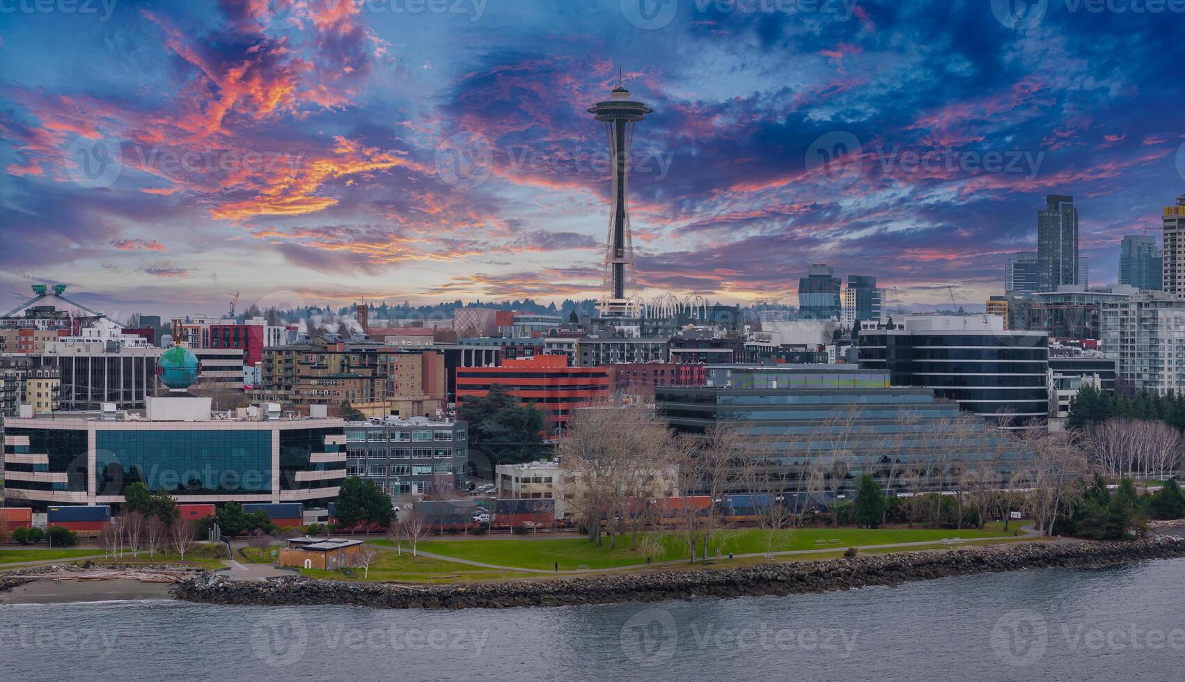 Downtown Seattle skyline featuring the Space Needle, modern skyscrapers, a vibrant sunset sky, and a waterfront park with trees and rocky shoreline. photo