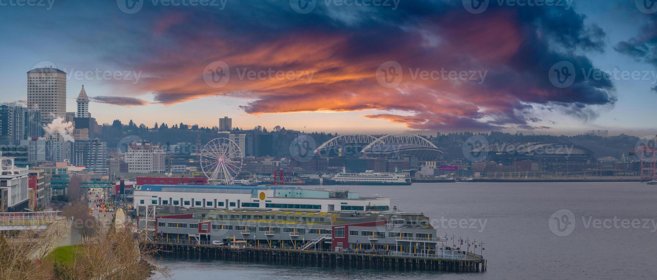 Aerial view of Seattle, Washington, featuring the Great Wheel, Smith Tower, Lumen Field, T Mobile Park, a pier, a cargo ship, and a vibrant sky. photo