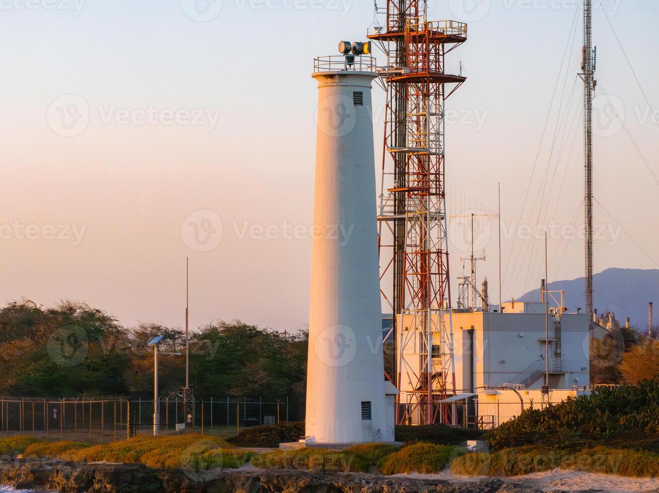 A white lighthouse near a red and white communication tower, surrounded by low vegetation and fenced area, with mountains in the background at sunset. photo
