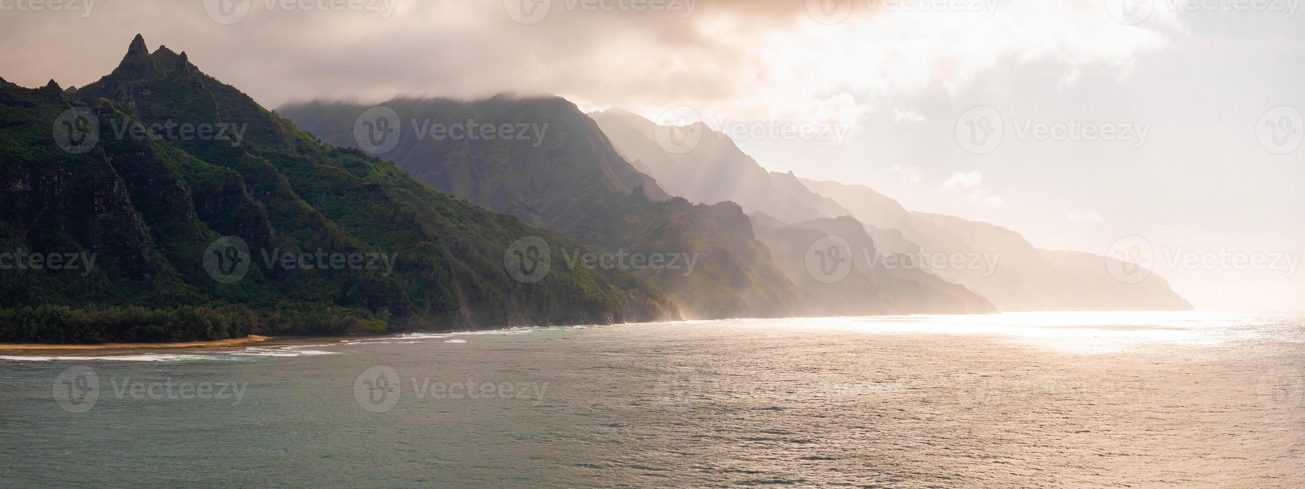 Aerial view of Kauai's Napali Coast with jagged green cliffs, a sandy beach, and turquoise Pacific waters under soft sunlight and scattered clouds. photo