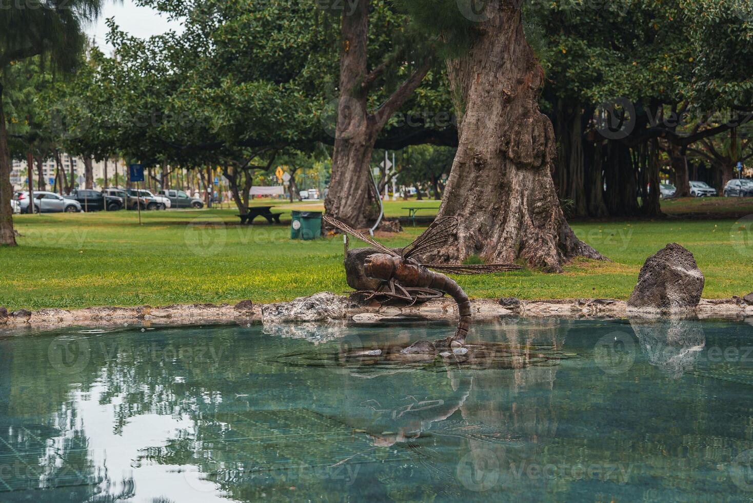 A tranquil park features a reflective pond with a metal fish sculpture, surrounded by trees, grass, benches, and a few parked cars in Hawaii. photo
