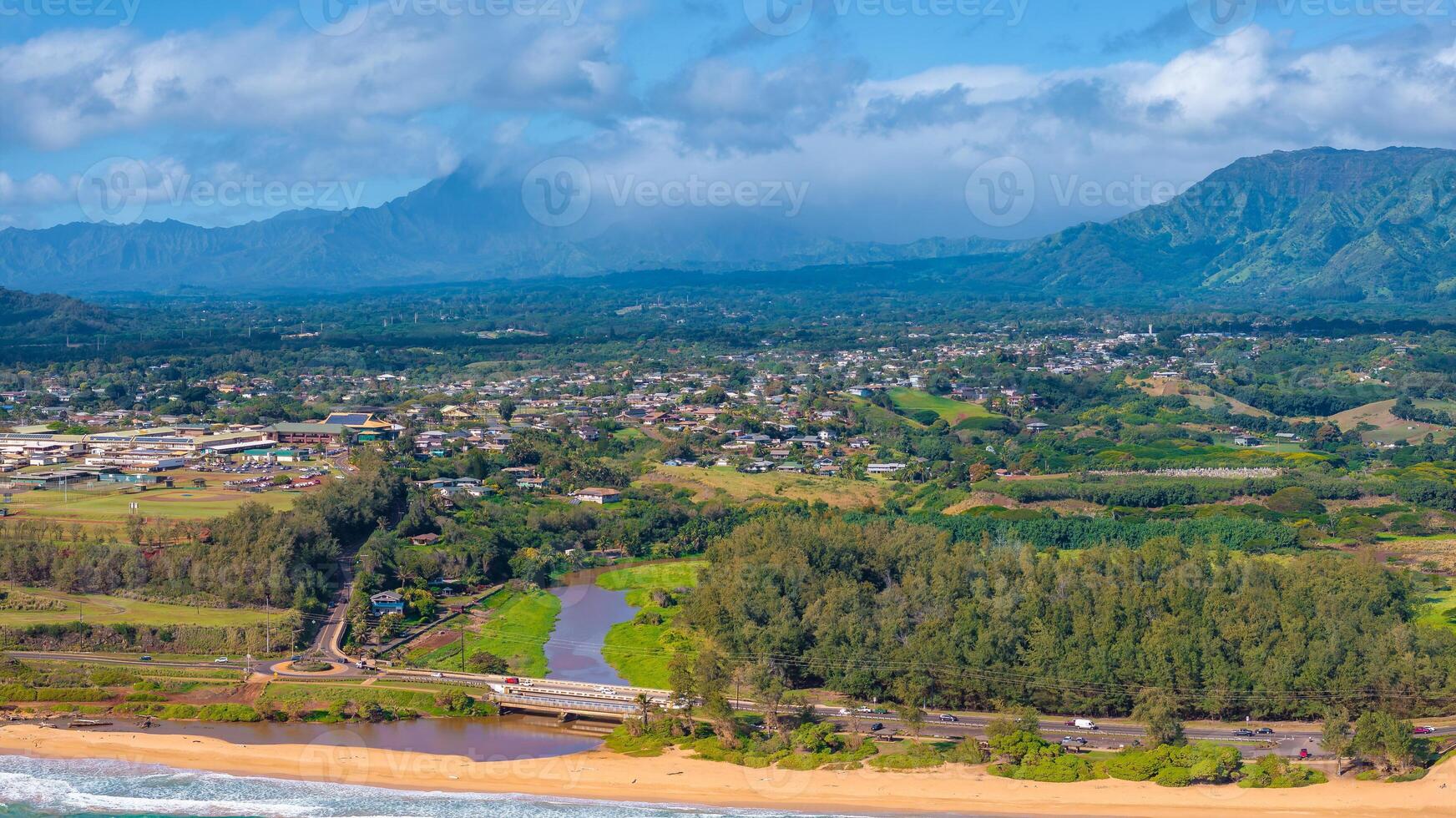 Aerial image of Kauai island, Hawaii, showing a sandy beach, winding river with a bridge, a town, verdant fields, and the Napali cliffs in the background. photo