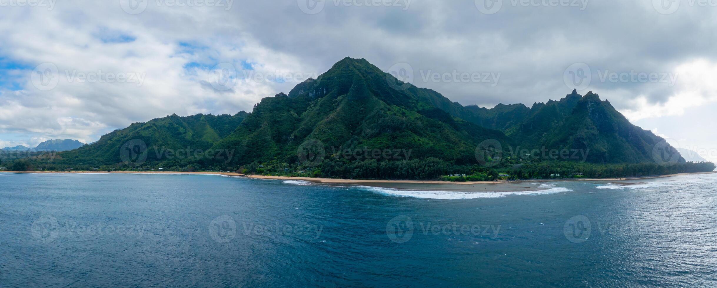 Aerial view of Kauai Island's Napali cliffs with lush greenery, sandy beach, ocean waves, rugged peaks, and a partly cloudy sky. photo